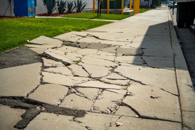 Damaged sidewalk with uneven and cracked concrete slabs caused by ground settlement.